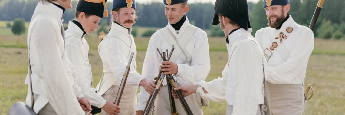 A group of historical reenactors in military uniforms stacking rifles outdoors in a field.
